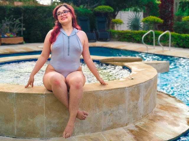 AlanisSweet, a chubby woman with red hair and glasses, sits on a pool edge wearing a gray, sleeveless, zip-up one-piece, showing her large, round tits and thick thighs.