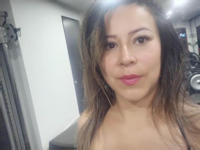 Close-up of Contelaya with medium brown skin, wavy brown hair, and pink lipstick. She has dark brown eyes and a slight smile. The image is brightly lit from above, highlighting her face. Contelaya is topless, with bare shoulders visible. The background is a dimly lit room with a door and gym equipment. The focus is on Contelaya's face. Her hair is slightly messy. The overall composition is simple and direct.