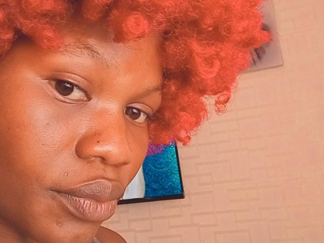 BelNetty with bright red curly hair, dark brown skin, and a small nose piercing, looking slightly to the side with a neutral expression. Her ass is not visible.