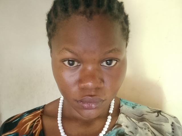 BelNetty has dark brown skin, small nose, and pouty lips with a slight tattoo under her chin. She wears a white bead necklace and a colorful patterned top.