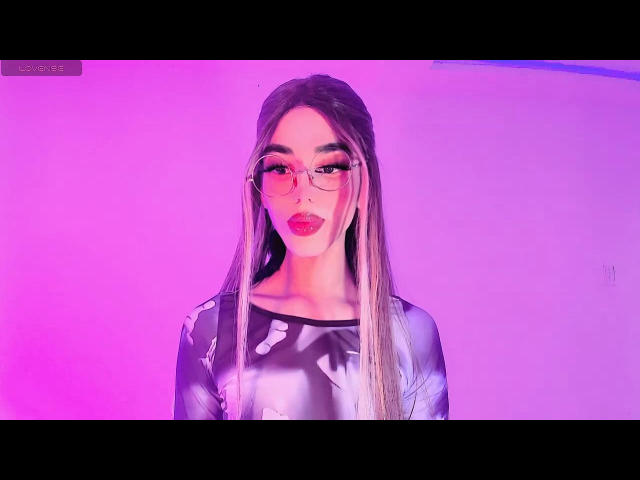 Aalanax, avec de longs cheveux blonds, des lunettes roses et du rouge à lèvres rouge, portant un haut noir brillant, avec un éclairage néon rose et violet. Les seins ne sont pas visibles, l'accent étant mis sur le visage et le haut du corps..