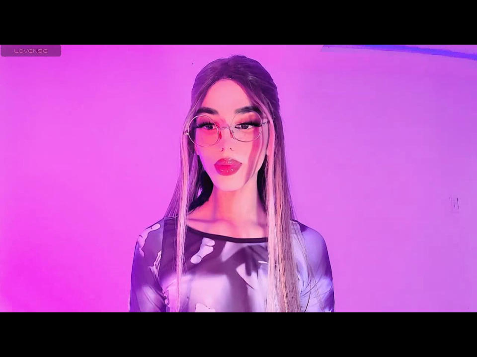 Eine Frau namens Aalanax mit langen blonden Haaren, rosa Brille und rotem Lippenstift, trägt ein glänzendes schwarzes Oberteil. Rosa und violette Neonlichter. Die Brust ist nicht sichtbar, der Fokus liegt auf dem Gesicht und dem Oberkörper..