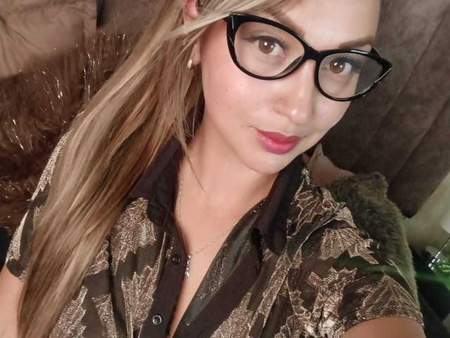 MaiyaJonesy with glasses, blonde hair, and pink lipstick, wearing a brown patterned shirt, showing a cute smile, and a small necklace, looking into the camera.
