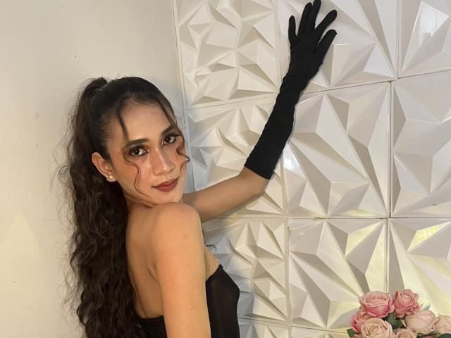 PreciousHorny with long curly brown hair, wearing black gloves and strapless black top, touching geometric white wall, smiling at camera, pink roses in bottom right corner.