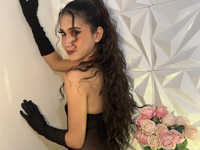 PreciousHorny with long curly brown hair, black gloves, and strapless black mesh dress, touching white wall, looking over shoulder, pink roses in bottom right.
