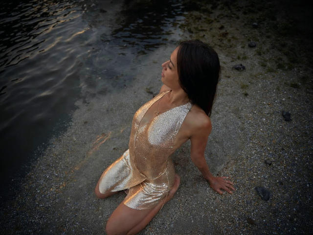 MaryLoveX kneels on gravel, wearing a shiny gold dress that clings to her tits and ass, with dark wet hair, gazing upwards, by a water's edge.
