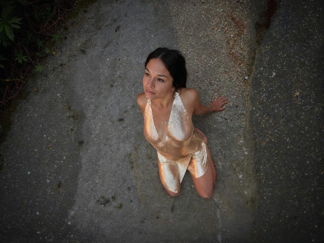 MaryLoveX on her knees, wearing a shiny, gold, halter dress, looking up with a cute, subtle smile. Her ass is slightly visible, dark hair, soft light.