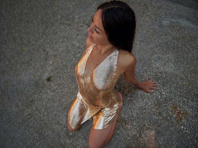 MaryLoveX with shiny gold dress, sitting on gravel, looking up, dark wet hair, small tits, slim, no pants, gold dress reflecting light, arms resting on gravel, smiling.