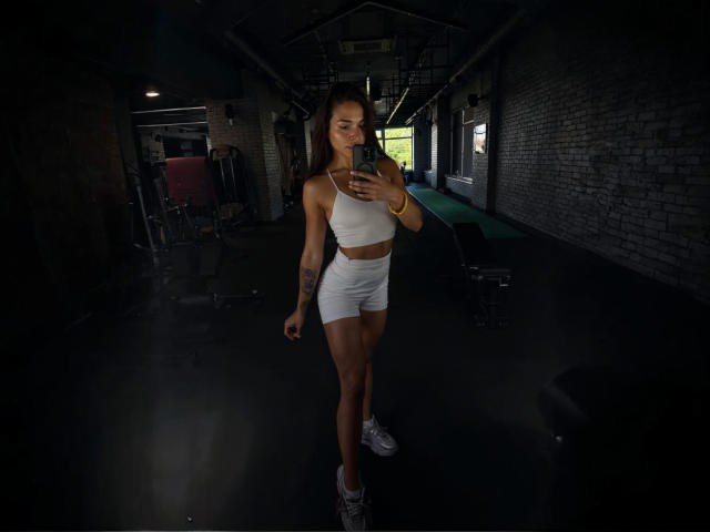 MaryLoveX, with a toned body and small tits, in a white sports bra and shorts, taking a selfie in a dark gym, wearing white sneakers and a yellow bracelet.