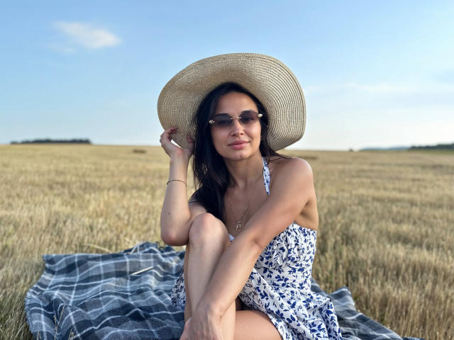 MaryLoveX sits in a field wearing a white floral dress, straw hat, and sunglasses, with her legs crossed, showing her tits and a cute smile. Blue checkered blanket.