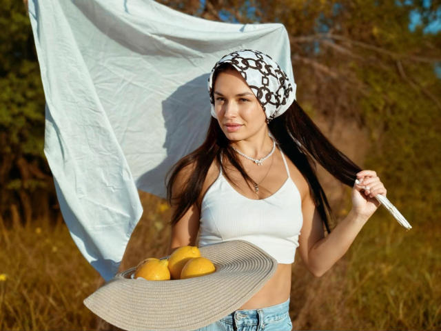 MaryLoveX with long brown hair, white crop top, and blue jeans, holding a sunhat and yellow lemons, wearing a black and white patterned headscarf, smiling.