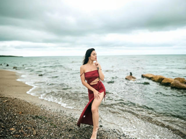 MaryLoveX with long black hair, wearing a red strapless dress, standing on a rocky beach, touching her chin, looking to the side, showing her bare ass and tits.