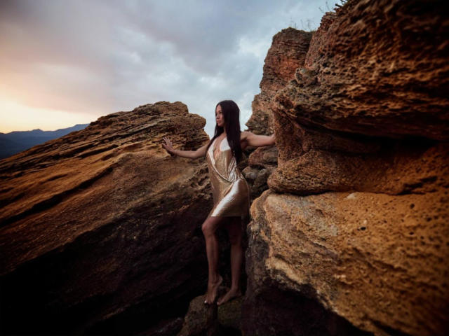 MaryLoveX stands between rocky cliffs, wearing a white dress with long necklace, arms out, brown hair, looking right, sunset in background, bare feet on rock, sexy, attractive.