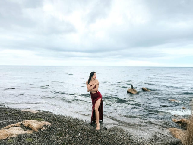 MaryLoveX stands on a rocky beach, arms crossed, wearing a red skirt and no top, showing her tits, with long black hair and a serious expression, cloudy sky.