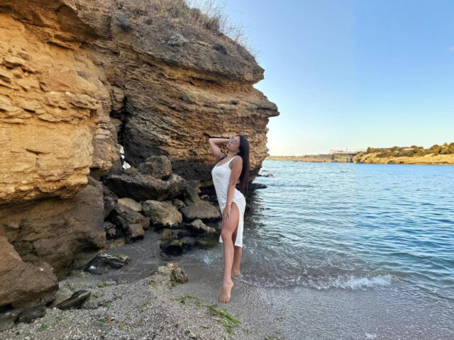 MaryLoveX with large tits in a white dress, standing by rocky shoreline, arms behind head, long dark hair, bare feet, water's edge, blue sky, sunlit, rocky texture.