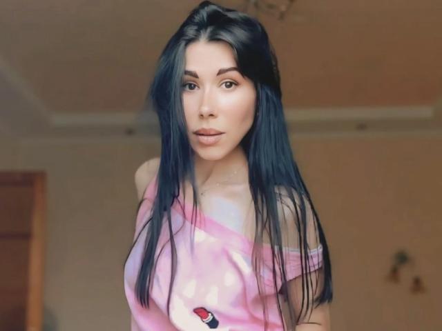 Glidia, avec ses longs cheveux noirs, son t-shirt rose à une épaule et un maquillage discret, dévoilant légèrement sa poitrine, regarde l'appareil photo avec une expression neutre..