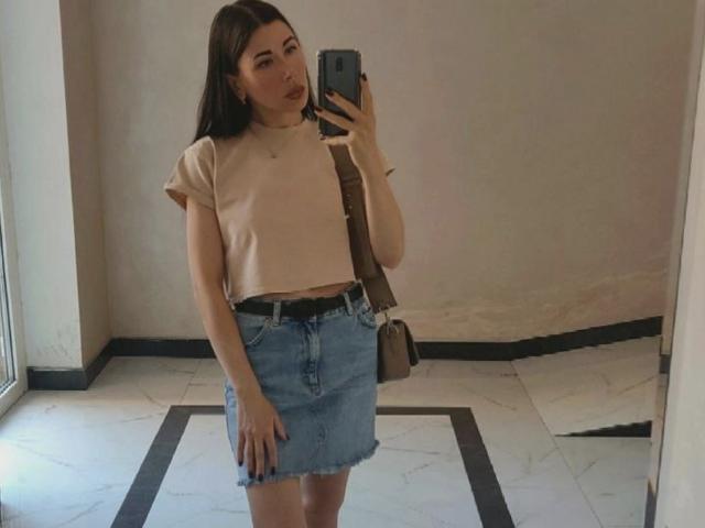 Glidia with long brown hair, tan tee, blue denim skirt, black belt, brown bag, holding phone, standing in mirror, showing her ass slightly. Reflection in mirror.