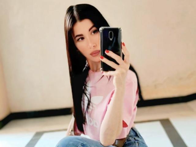 Glidia with long black hair, pink shirt, and red nails, holding a phone, taking a selfie, showing her face and upper body, sitting on a floor with striped tiles.