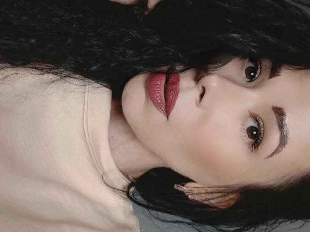 Close-up of Glidia with dark, wavy hair partially covering her face. She has light skin, brown eyes, and thick, well-groomed eyebrows. Glidia's lips are painted a bold red, contrasting with her fair complexion. She's wearing a beige top, and the image focuses on her facial expression, which is neutral yet slightly curious. The lighting is even, highlighting the texture of her hair and the subtle shine on her skin. The composition centers on her face, with her hair framing her features.