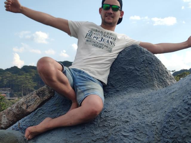 CharlieYeah with arms out, sitting on a large rock, wearing a white tee and blue shorts, green sunglasses, and a black cap, smiling with a slight mustache. Barefoot.