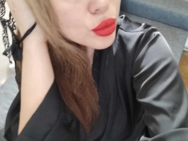 SofiaBells with red lipstick, black satin shirt, and long brown hair, pouting her glossy red lips, showing her bare shoulder and black lace strap.