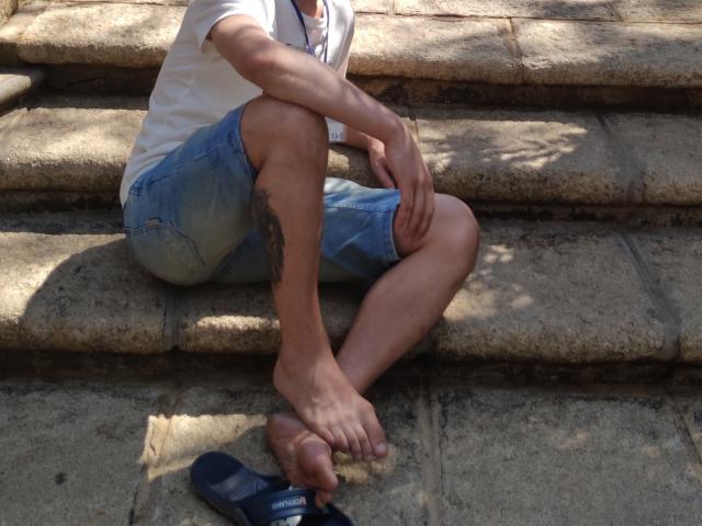 CharlieYeah is sitting on stone steps, wearing a white tee and blue shorts, with a tattoo on his right leg, barefoot, and his navy flip-flops on the ground.