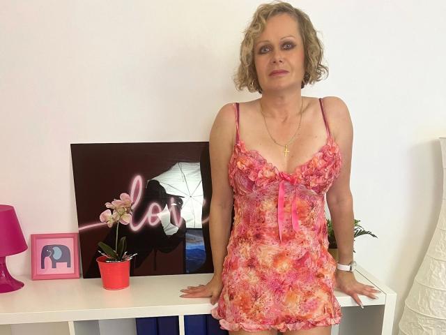 KatharinaSky-hot stands in a pink floral nightie, showing her tits and small ass. She has curly blonde hair, a gold cross necklace, and a white wristband.