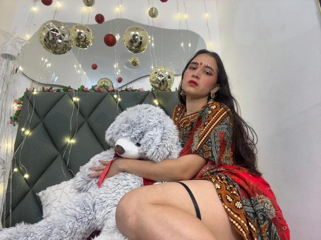JalimaMona with red dress, patterned fabric, holding fluffy white teddy bear, red ribbon, black garter, dark hair, red makeup, sitting on green headboard, string lights, gold balloons.