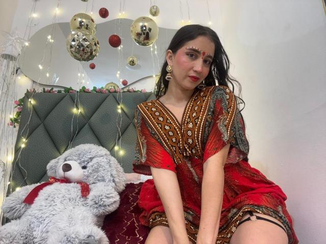 JalimaMona with dark wet hair, red and brown patterned dress, facial jewelry, and red lipstick, sitting on bed with gray stuffed teddy and string lights. Boobs visible under dress.