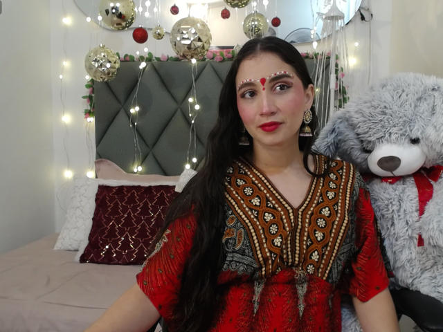 JalimaMona with long black hair, red dress, and intricate patterns, red makeup, and a red dot on her forehead, standing next to a fluffy gray teddy bear.