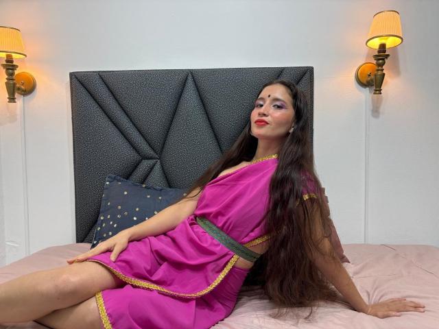 JalimaMona, avec de longs cheveux bruns, du rouge à lèvres rouge et du fard à paupières violet, portant une robe rose avec des détails jaunes, assise sur un lit, les jambes légèrement écartées, montrant ses fesses..