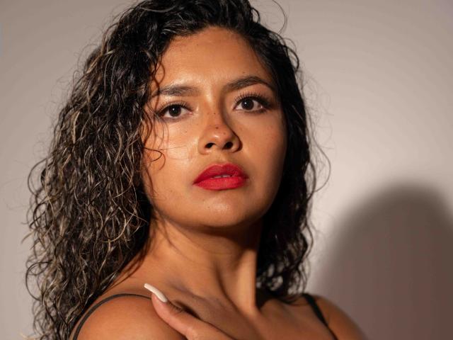 JedaZore tiene el cabello castaño, rizado y húmedo, ojos oscuros y un lápiz labial rojo brillante. Su piel bronceada y el maquillaje sutil resaltan su rostro atractivo. Lleva un top negro con tirantes..