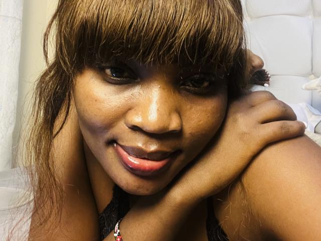 SexyEvaa with brown hair, bangs, and dark skin, tongue slightly out, looking at camera, resting cheek on hand, wearing black lace, in a bedroom with white furniture.