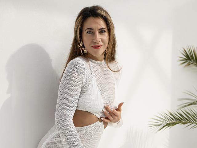 KateShanny with long brown hair, white mesh top, exposing her tits, and matching white bottom, holding her top, standing against a white wall, palm leaves in the background.