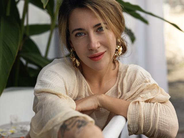KateShanny with light brown hair, green eyes, and gold earrings, wearing a beige, waffle-texture top, leaning on a white surface, smiling softly, tattoo on right forearm.
