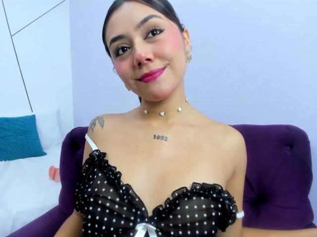 AlannaJonson with dark hair, pink lipstick, and a black polka dot bra with ruffles, showing her small tits and a tattoo on her right shoulder. Seated on a purple chair.