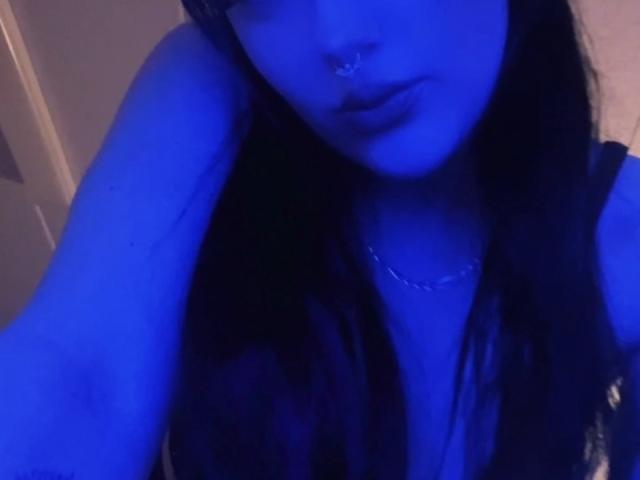 Blue-lit selfie of ClaireSage with long black hair, nose ring, and silver necklace, showing her pouty lips and bare shoulders, focusing on her chest and upper body.