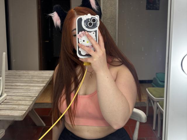 VicluX with cat ears, pink sports bra, and yellow earbuds, taking a mirror selfie. Long red hair, phone with cat face case, white table, and wooden floor.