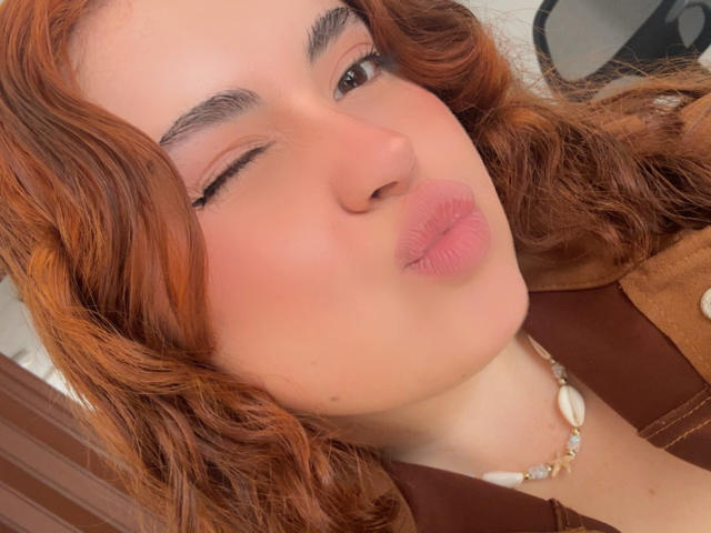 VicluX with wavy red hair, one eye closed, puckered lips, wearing a brown top and shell necklace, has a light skin tone and subtle makeup.