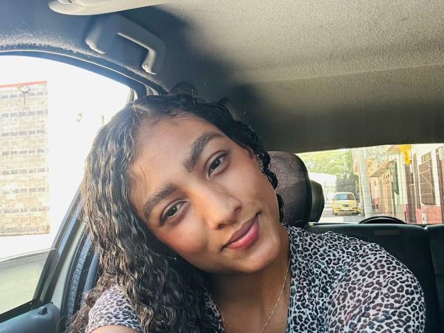 NatalyBlonder with curly black hair, brown skin, and a leopard print shirt, leaning into a car seat, smiling softly, with a subtle pink lip and a cute nose.