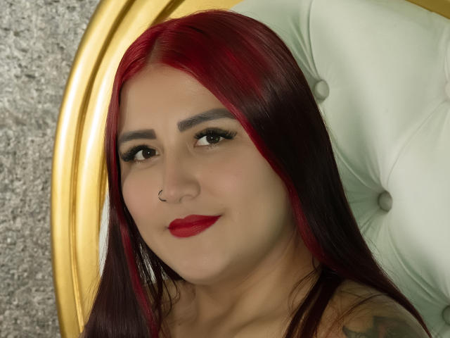 MarianaRamos has red and black dyed hair, a nose ring, red lipstick, and is smiling with dark eyeliner and tattoos visible on her shoulder. She's seated in a green chair.