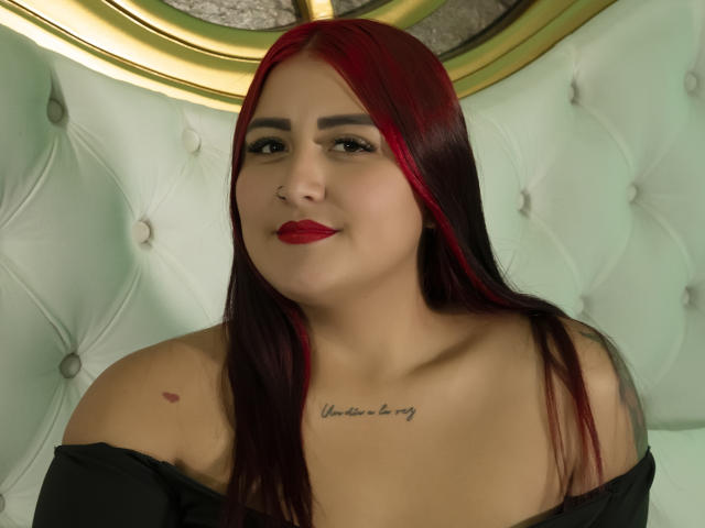 MarianaRamos with red and black hair, red lipstick, black off-shoulder top, tattoo under her tits, smiling at the camera. Her tits are small, and she has a cute face.
