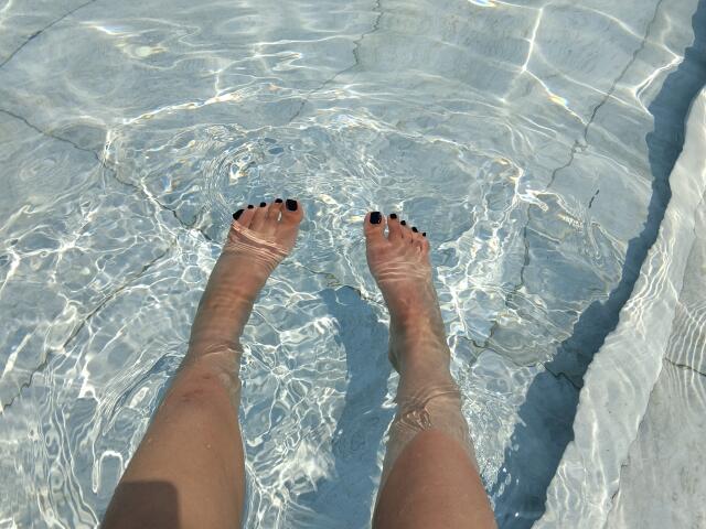 I piedi di Mllevv nell'acqua limpida, le dita dei piedi con lo smalto nero, le gambe leggermente abbronzate, riflessi luminosi sulla superficie dell'acqua, il fondo della piscina visibile..