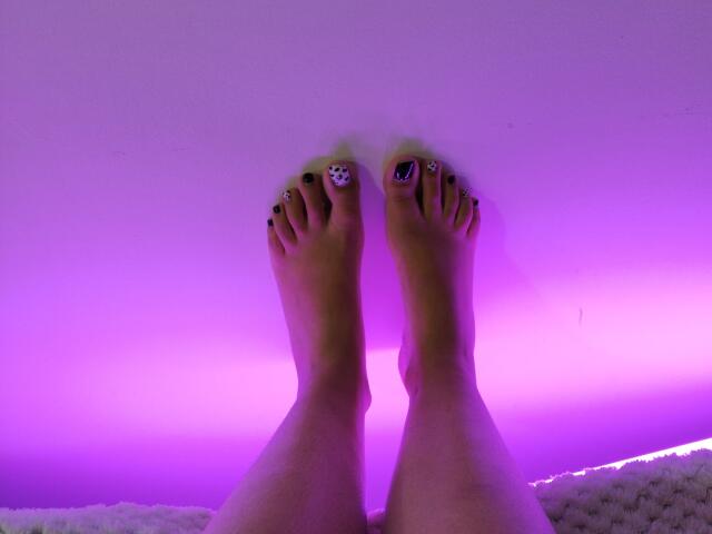 Mllevv's feet with purple-tinted skin, toes painted green and black with decorative nail art, legs slightly parted, resting on a white textured surface.