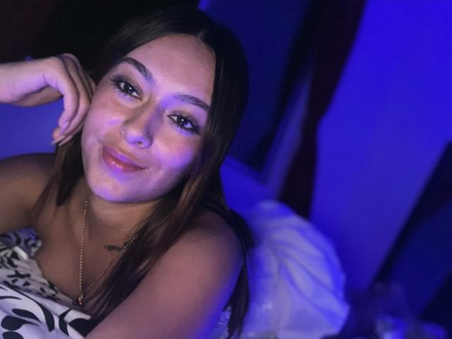 DamiAmbani has long brown hair, dark eyebrows, and a cute smile with a small nose piercing. She's topless with a white and black pillow, under blue lighting.