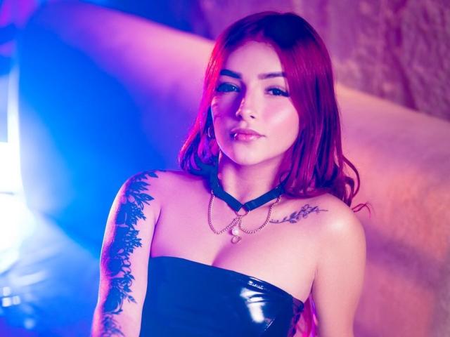 ErotikScarlett with red hair, black strapless latex top, tattoos on left arm, choker with chain, looking at camera, blue and pink lighting, sexy, confident, pouty lips.