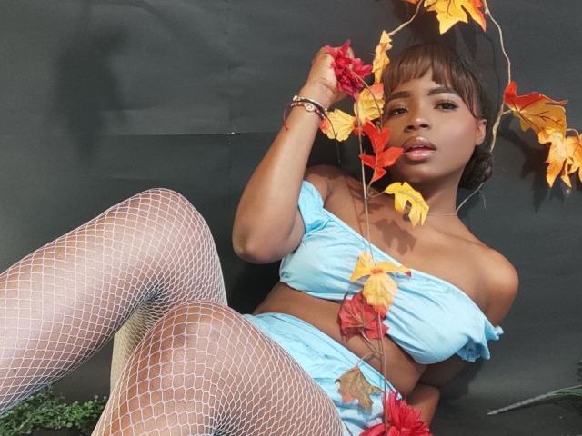 LindaBird with brown skin, small tits, wearing a blue strapless top and matching shorts, fishnet stockings, and autumn leaves in her hair, lying down, looking at the camera.