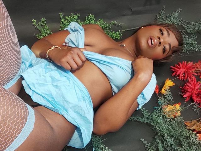 LindaBird is lying on her back, wearing a blue dress pulled up to expose her tits, white fishnet stockings, and a gold bracelet. She has dark skin and a sexy smile.