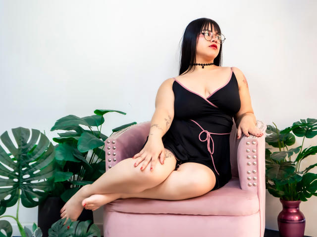 AnneStonee with black hair, glasses, choker, black dress, pink trim, sitting on pink chair, legs crossed, showing her ass, tattoo on arm, plants around, red nails, looking up.