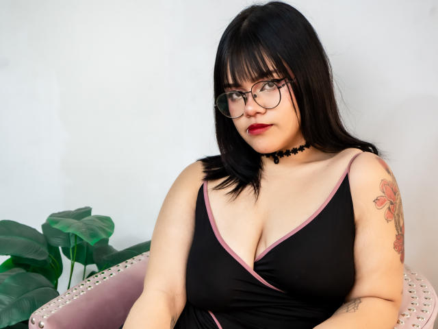 AnneStonee with black hair, glasses, and red lipstick, wearing a black dress with pink trim, showing her big tits, choker, and floral tattoo on her right arm.